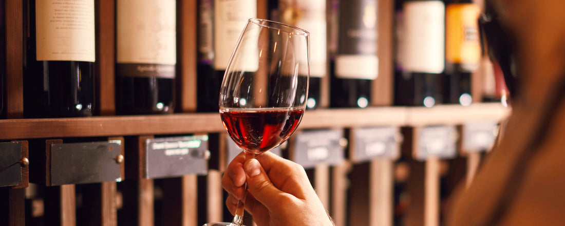 Glas Rotwein gehalten von einer Hand vor einem Regal mit verschwommenen Weinflaschen